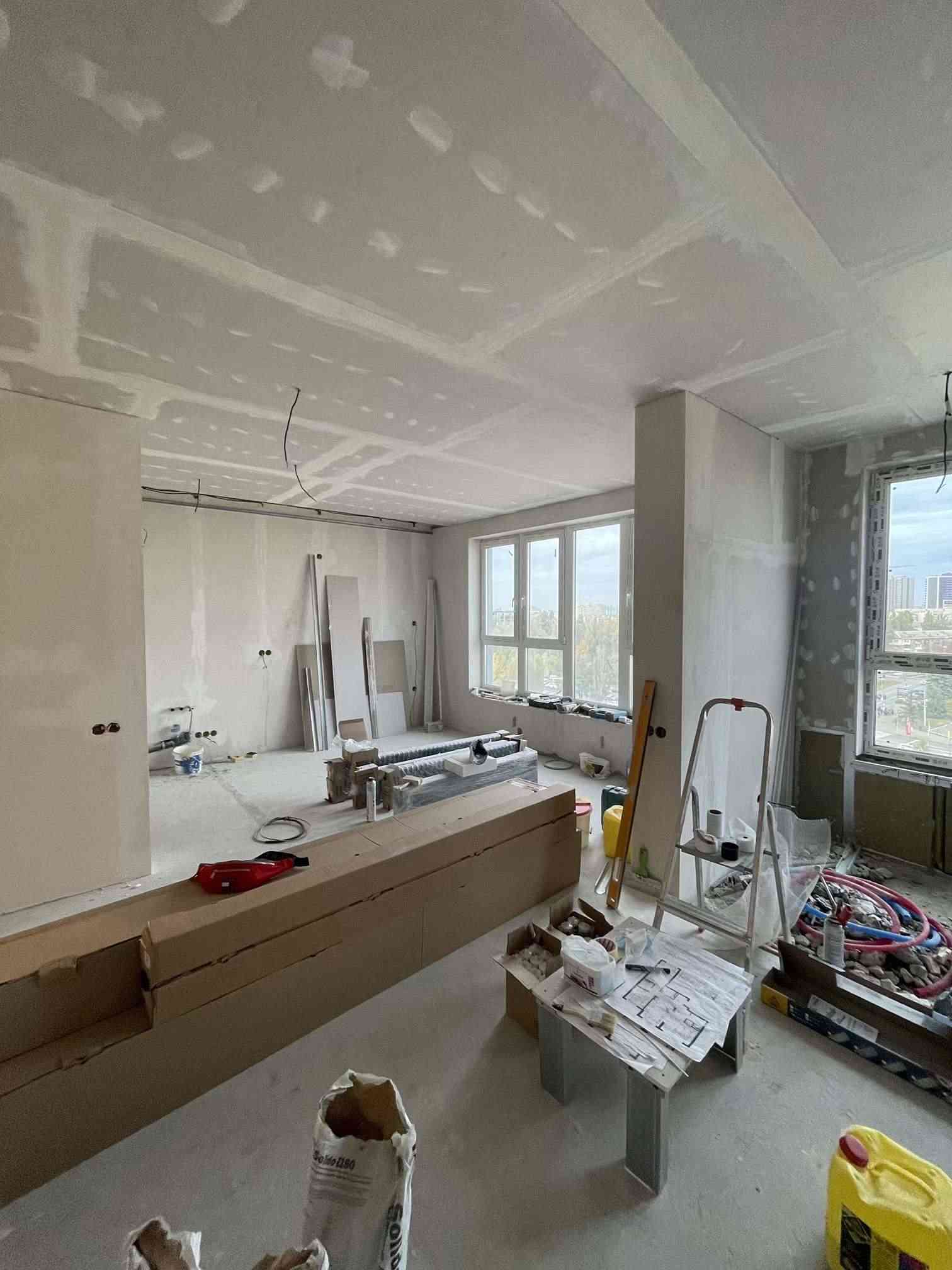 Open-plan apartment renovation in progress with drywall ceiling, materials on site, and large windows, Rimmart project